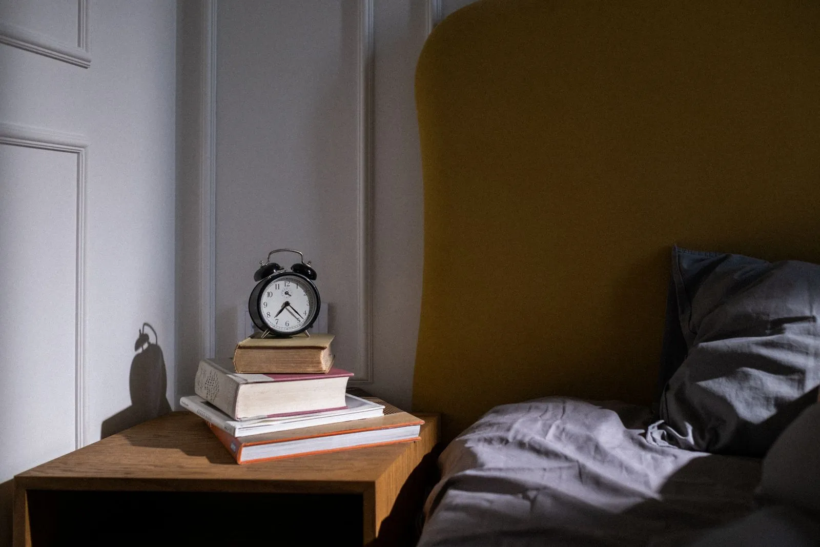 Black twin-bell alarm clock on a stack of books on a wooden nightstand beside an unmade bed