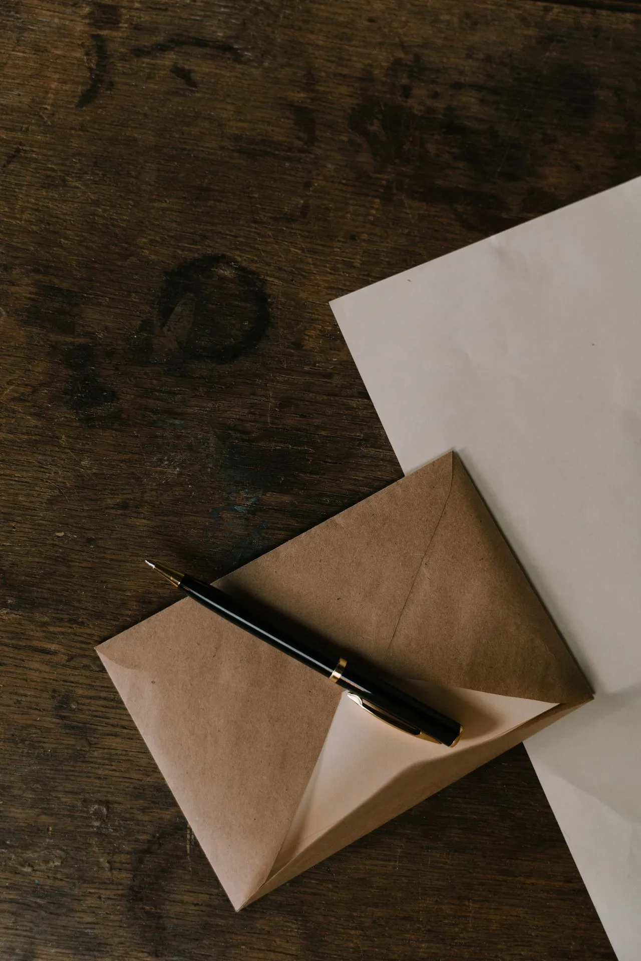 Black fountain pen resting on a kraft paper envelope and a sheet of letter paper on a dark wooden desk