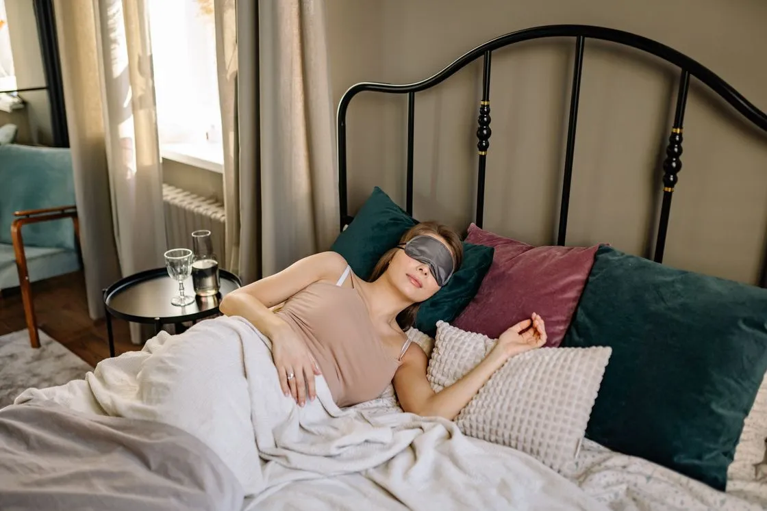 Woman lying in bed in a gray eye mask among teal and plum cushions