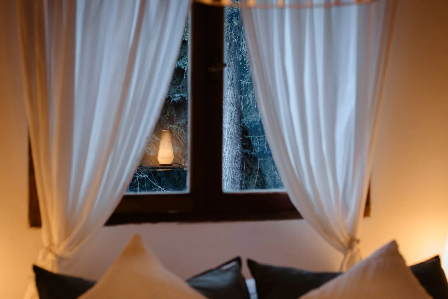 White curtains framing a window onto frosted trees at dusk, warm lamp glow
