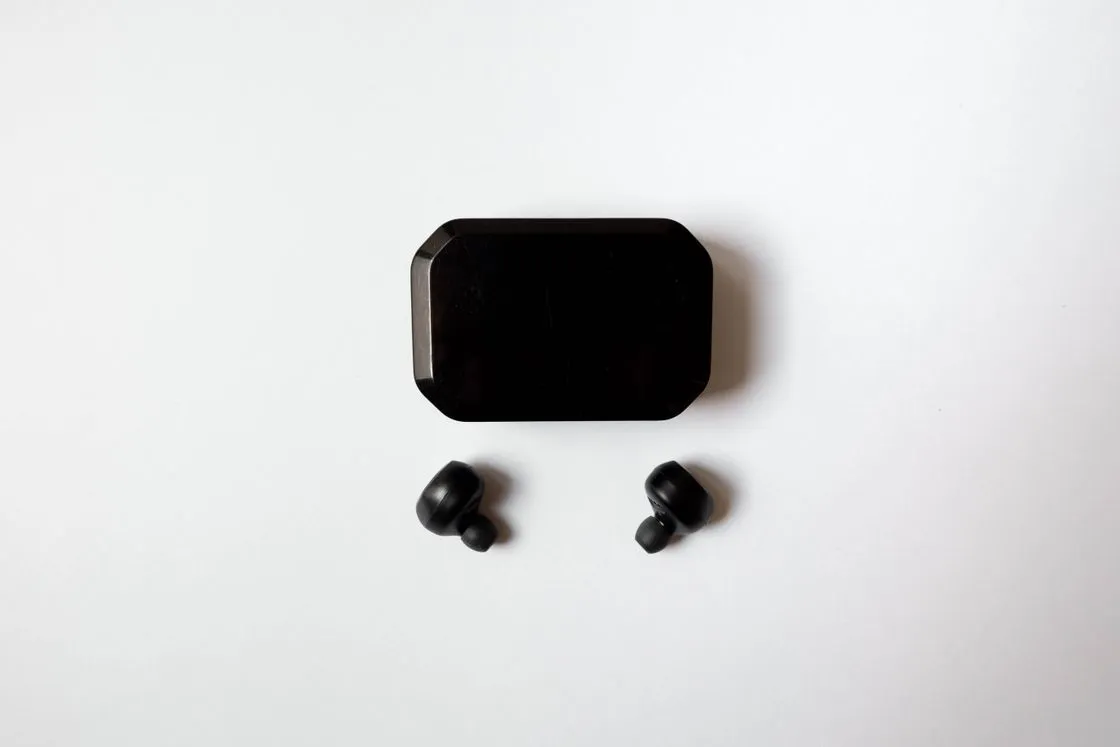 Two small black wireless earbuds beside their closed charging case on a white surface
