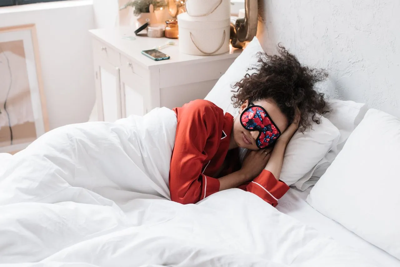 Woman in red pajamas asleep on her side under a white duvet wearing a patterned eye mask